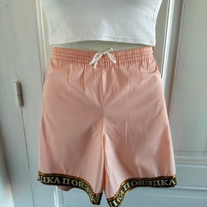 Puede incluir: Shorts deportivos de color rosa con un ribete marr&oacute;n y dorado que dice "ПІКА ПОЕЗДКА".