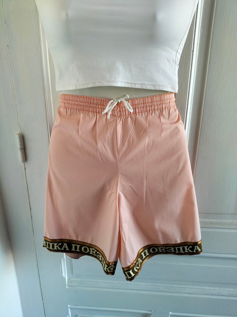 Puede incluir: Shorts deportivos de color rosa con un ribete marr&oacute;n y dorado que dice "ПІКА ПОЕЗДКА".