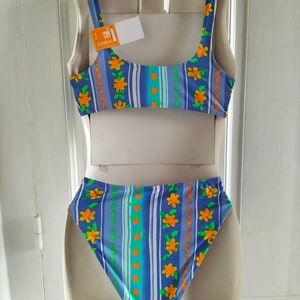 Op de afbeelding: Blauw-wit gestreepte bikini met een bloemenpatroon. De top heeft een ronde hals en dunne bandjes. De onderkant is hoog getailleerd.