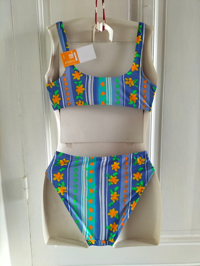 Op de afbeelding: Blauw-wit gestreepte bikini met een bloemenpatroon. De top heeft een ronde hals en dunne bandjes. De onderkant is hoog getailleerd.