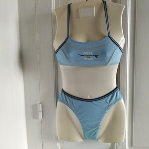 Op de afbeelding: Lichtblauwe bikiniset op een mannequin. De top heeft dunne bandjes en de tekst "OCEAN SURF WEAR". Donkerblauwe biezen omlijnen de randen. De bijpassende broek maakt de tweedelige badkleding compleet.