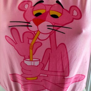Pode incluir: Body rosa com uma imagem da Pantera Cor-de-Rosa bebendo com um canudo. A Pantera Cor-de-Rosa tem olhos amarelos e nariz vermelho. O body tem decote redondo e acabamento brilhante. O texto "&copy; 1965 UNITED ARTISTS CORPORATION" &eacute; vis&iacute;vel.