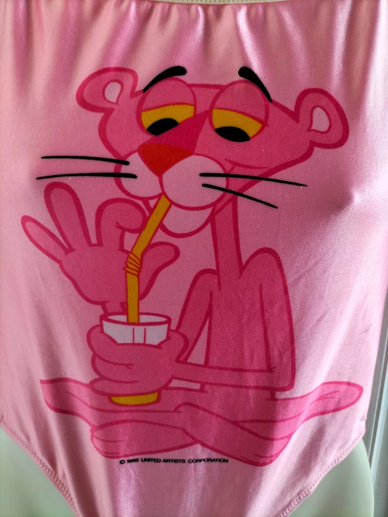 Pode incluir: Body rosa com uma imagem da Pantera Cor-de-Rosa bebendo com um canudo. A Pantera Cor-de-Rosa tem olhos amarelos e nariz vermelho. O body tem decote redondo e acabamento brilhante. O texto "&copy; 1965 UNITED ARTISTS CORPORATION" &eacute; vis&iacute;vel.