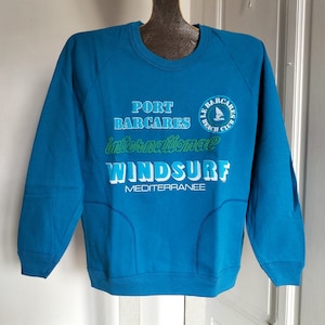 Può includere: Felpa blu turchese con la scritta "PORT BARCARES international WINDSURF MEDITERRANEE" in bianco e verde. Un logo circolare con una barca a vela è in alto a destra. La felpa ha girocollo e maniche a raglan.