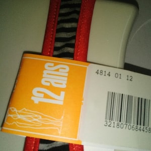 May include: Orange tag with the text "12 ans" and a white outline. The tag is attached to a red and black striped fabric with a white background. The tag also has a barcode with the number 3218070684458.