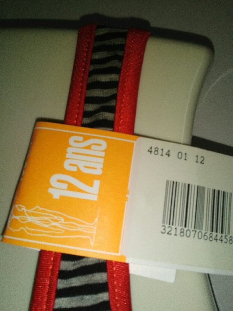 May include: Orange tag with the text "12 ans" and a white outline. The tag is attached to a red and black striped fabric with a white background. The tag also has a barcode with the number 3218070684458.