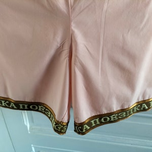 Puede incluir: Pantalones cortos de color rosa con ribete marr&oacute;n y dorado con el texto "КА ПОЕЗДКА" en escritura cir&iacute;lica.