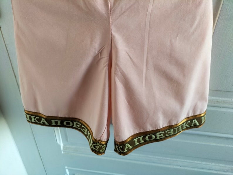 Puede incluir: Pantalones cortos de color rosa con ribete marr&oacute;n y dorado con el texto "КА ПОЕЗДКА" en escritura cir&iacute;lica.