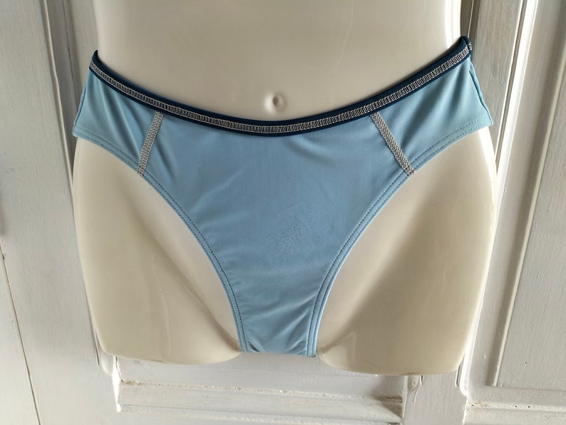 Op de afbeelding: Lichtblau ondergoed met een donkerblauwe rand en decoratieve stiksels. Het ondergoed wordt op een mannequin getoond. Het kledingstuk heeft een lage taille en een gladde, zijdeachtige textuur.