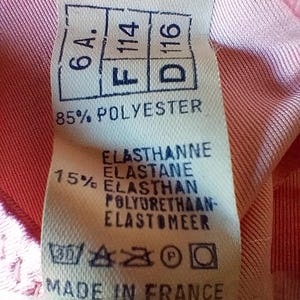 Pode incluir: Um close-up de uma etiqueta de roupa com o texto "6A. F114 D116" e "85% POLYESTER". A etiqueta tamb&eacute;m inclui instru&ccedil;&otilde;es de lavagem e o texto "MADE IN FRANCE". A etiqueta est&aacute; em um tecido rosa.