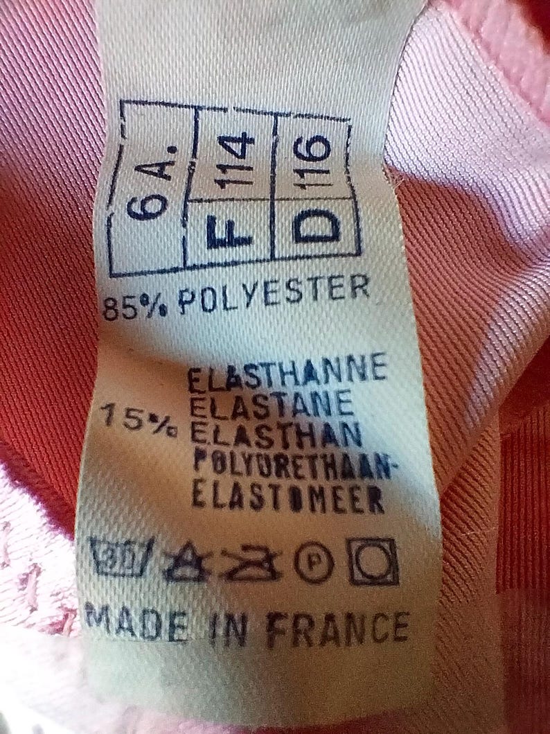 Pode incluir: Um close-up de uma etiqueta de roupa com o texto "6A. F114 D116" e "85% POLYESTER". A etiqueta tamb&eacute;m inclui instru&ccedil;&otilde;es de lavagem e o texto "MADE IN FRANCE". A etiqueta est&aacute; em um tecido rosa.