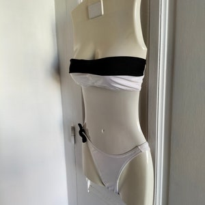 K&ouml;nnte beinhalten: Ein wei&szlig;er und schwarzer Bikini-Oberteil und -Unterteil an einer wei&szlig;en Schaufensterpuppe. Das Oberteil ist ein Bandeau-Stil mit einem schwarzen Band und das Unterteil ist ein hochgeschnittener Stil mit einer schwarzen Schleife.