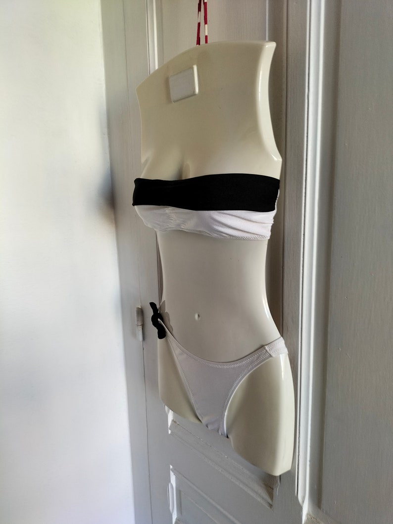 K&ouml;nnte beinhalten: Ein wei&szlig;er und schwarzer Bikini-Oberteil und -Unterteil an einer wei&szlig;en Schaufensterpuppe. Das Oberteil ist ein Bandeau-Stil mit einem schwarzen Band und das Unterteil ist ein hochgeschnittener Stil mit einer schwarzen Schleife.