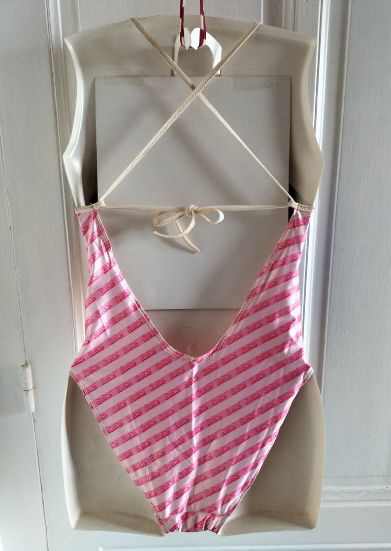 Puede incluir: Traje de ba&ntilde;o de rayas rosa y blanco con espalda baja y tirantes finos. El traje de ba&ntilde;o tiene un peque&ntilde;o logotipo repetido en toda la tela.