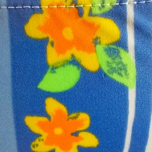 Op de afbeelding: Close-up van een blauwe stof met een witte streep en een herhalend patroon van oranje en gele bloemen met groene bladeren.