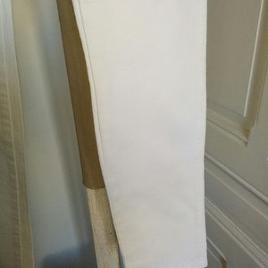 May include: A pair of white pants hanging on a wooden rack. The pants are made of a smooth, lightweight fabric.