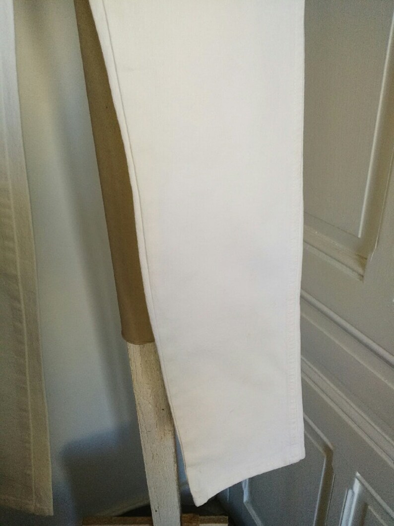 May include: A pair of white pants hanging on a wooden rack. The pants are made of a smooth, lightweight fabric.