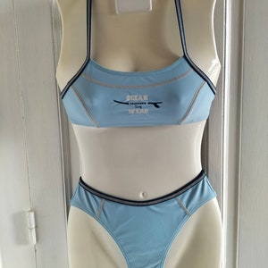 Op de afbeelding: Lichtblauw tweedelig badpak op een mannequin. De top heeft dunne bandjes en de tekst "OCEAN CALIFORNIA SURF WEAR". Donkerblauwe biezen omlijnen de randen. De onderkant is een bijpassende bikinibroek.