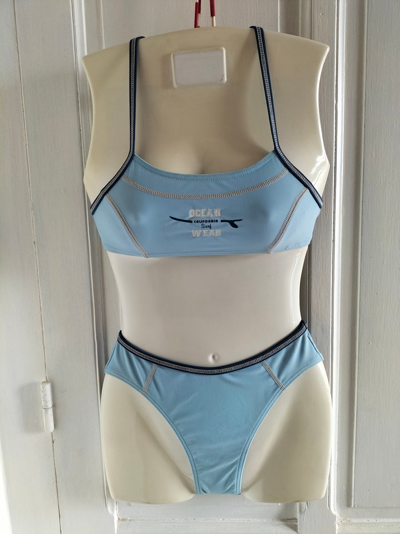 Op de afbeelding: Lichtblauw tweedelig badpak op een mannequin. De top heeft dunne bandjes en de tekst "OCEAN CALIFORNIA SURF WEAR". Donkerblauwe biezen omlijnen de randen. De onderkant is een bijpassende bikinibroek.