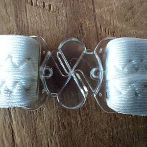 K&ouml;nnte beinhalten: Wei&szlig;e elastische B&auml;nder mit einer klaren, verstellbaren Kunststoffschnalle. Die Schnalle hat ein vierseitiges Design.
