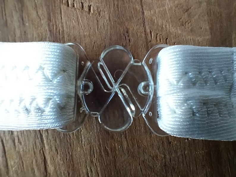 K&ouml;nnte beinhalten: Wei&szlig;e elastische B&auml;nder mit einer klaren, verstellbaren Kunststoffschnalle. Die Schnalle hat ein vierseitiges Design.