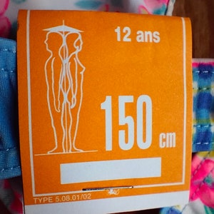 Puede incluir: Una etiqueta naranja con el texto "12 ans" y "150 cm" est&aacute; adherida a una prenda con estampado floral. La etiqueta tambi&eacute;n presenta una ilustraci&oacute;n estilizada de dos figuras y el texto "TYPE 5.08.01/02".