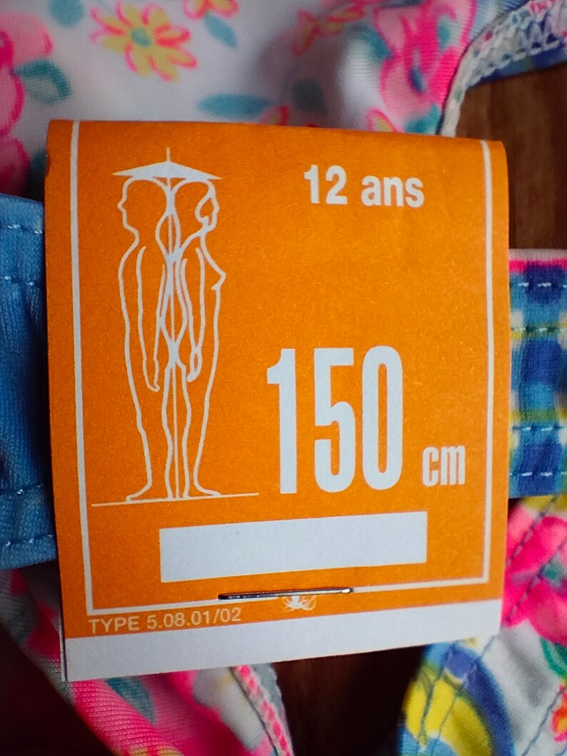 Puede incluir: Una etiqueta naranja con el texto "12 ans" y "150 cm" est&aacute; adherida a una prenda con estampado floral. La etiqueta tambi&eacute;n presenta una ilustraci&oacute;n estilizada de dos figuras y el texto "TYPE 5.08.01/02".