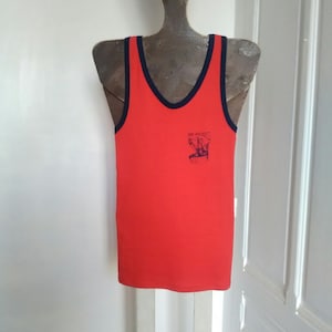 May include: A red vest top with navy blue trim around the neckline and armholes. The vest top features a small graphic and text on the front that reads "Port Macades". It is displayed on a metal mannequin.