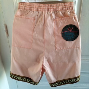 Puede incluir: Shorts de color melocot&oacute;n con un parche blanco y negro que dice "Laguna Beach" en la parte posterior. Los pantalones cortos tienen un ribete marr&oacute;n y dorado con la palabra "поездка" escrita en el ribete.