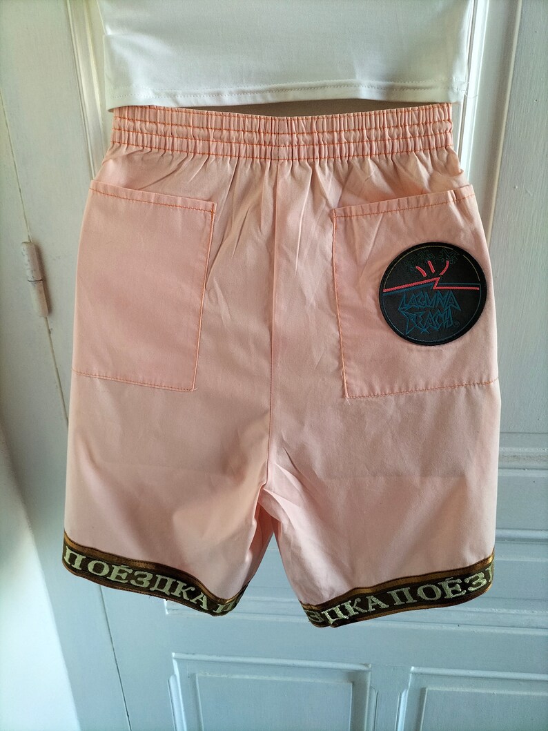 Puede incluir: Shorts de color melocot&oacute;n con un parche blanco y negro que dice "Laguna Beach" en la parte posterior. Los pantalones cortos tienen un ribete marr&oacute;n y dorado con la palabra "поездка" escrita en el ribete.