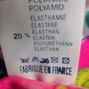 Puede incluir: Una etiqueta de tela blanca con texto en azul y negro, incluyendo "POLYAMIDE", "ELASTHANNE" y "FABRIQUE EN FRANCE". La etiqueta es parcialmente visible contra una tela rosa con estampado floral.