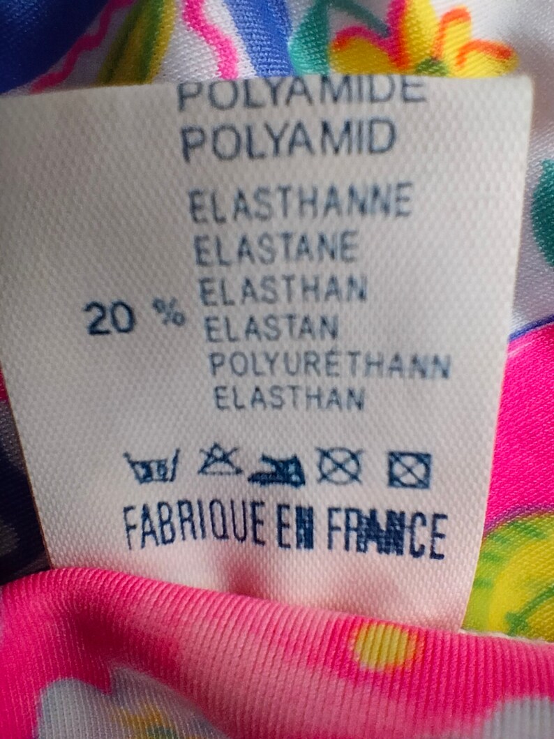 Puede incluir: Una etiqueta de tela blanca con texto en azul y negro, incluyendo "POLYAMIDE", "ELASTHANNE" y "FABRIQUE EN FRANCE". La etiqueta es parcialmente visible contra una tela rosa con estampado floral.