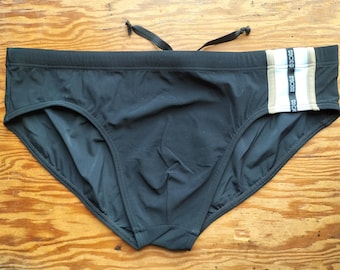 Puede incluir: Bañador tipo slip negro con cordón en la cintura y un panel lateral. El panel lateral consta de una franja blanca flanqueada por dos franjas de color beige, con la palabra "HOM" impresa en negro sobre la franja blanca central.