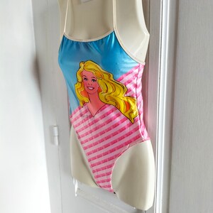 Puede incluir: Un ba&ntilde;ador rosa y azul con un estampado gr&aacute;fico de Barbie. El ba&ntilde;ador tiene tirantes finos y un patr&oacute;n a rayas.