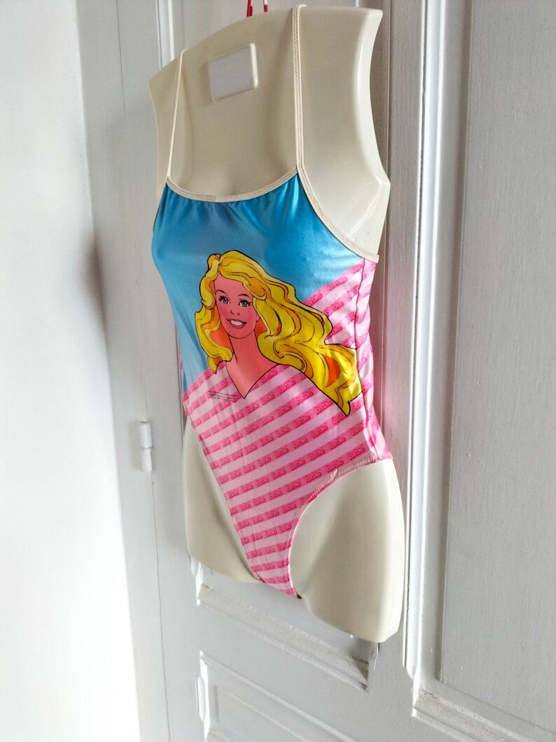 Puede incluir: Un ba&ntilde;ador rosa y azul con un estampado gr&aacute;fico de Barbie. El ba&ntilde;ador tiene tirantes finos y un patr&oacute;n a rayas.