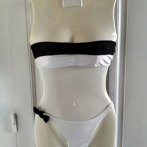 K&ouml;nnte beinhalten: Wei&szlig;er Bikini mit einem schwarzen Bandeau-Oberteil und einem wei&szlig;en Unterteil mit einer schwarzen Schleife.