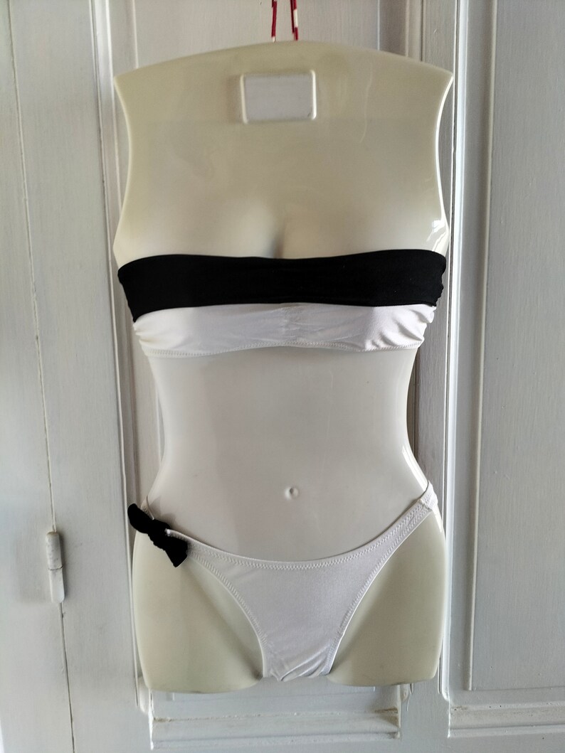 K&ouml;nnte beinhalten: Wei&szlig;er Bikini mit einem schwarzen Bandeau-Oberteil und einem wei&szlig;en Unterteil mit einer schwarzen Schleife.