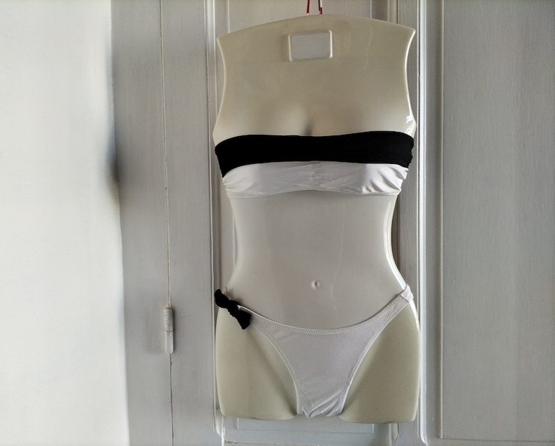 K&ouml;nnte beinhalten: Wei&szlig;er, tr&auml;gerloser Bikini-Oberteil mit schwarzem Band und einem passenden wei&szlig;en Unterteil mit schwarzer Schleife.