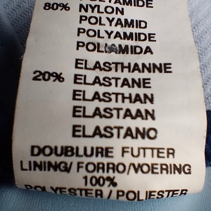 Op de afbeelding: Een wit kledinglabel met zwarte tekst met details over de stof: 80% Polyamide, 20% Elastaan en 100% Polyester. Het label vermeldt ook de voering en vertalingen van het woord "voering".