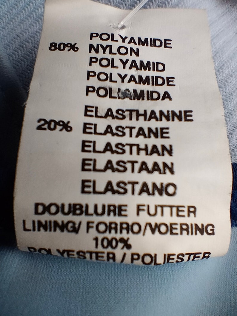 Op de afbeelding: Een wit kledinglabel met zwarte tekst met details over de stof: 80% Polyamide, 20% Elastaan en 100% Polyester. Het label vermeldt ook de voering en vertalingen van het woord "voering".