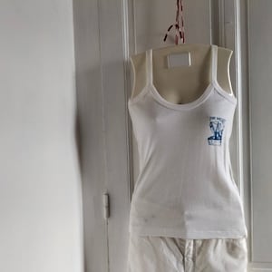 May include: White tank top with a blue graphic of a boat and the text "Port Yacht" on a white mannequin.