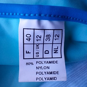 May include: A white tag with black text showing sizing information for a garment. The tag reads "F 40", "US/UK 12", "D 38", "NL 12". The tag also lists the fabric composition as 80% polyamide and 20% nylon.