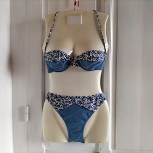 May include: A two-piece swimsuit displayed on a mannequin. The top is a blue, underwire bra with a floral patterned trim. The matching bottoms are high-waisted with a similar floral trim. The swimsuit is made of a denim-like material.