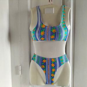 Op de afbeelding: Een blauw-wit gestreepte bikini met oranje bloemen. De bikini top heeft een ronde hals en de onderkant is een hoge snit.