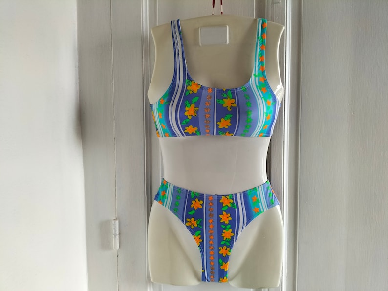 Op de afbeelding: Een blauw-wit gestreepte bikini met oranje bloemen. De bikini top heeft een ronde hals en de onderkant is een hoge snit.