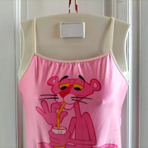 Pode incluir: Camisola rosa com um gr&aacute;fico da Pantera Cor-de-Rosa. A personagem de desenho animado &eacute; retratada bebendo de uma caneca com um canudo. A camisola tem al&ccedil;as finas e uma borda branca ao redor do decote. A roupa est&aacute; pendurada em um cabide branco.