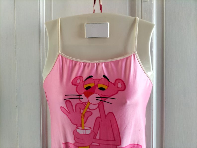 Pode incluir: Camisola rosa com um gr&aacute;fico da Pantera Cor-de-Rosa. A personagem de desenho animado &eacute; retratada bebendo de uma caneca com um canudo. A camisola tem al&ccedil;as finas e uma borda branca ao redor do decote. A roupa est&aacute; pendurada em um cabide branco.