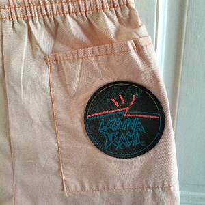 Puede incluir: Pantalones cortos de color melocot&oacute;n con un parche negro grande en el bolsillo trasero. El parche tiene un rayo blanco y el texto "Laguna Beach" en azul y rosa.