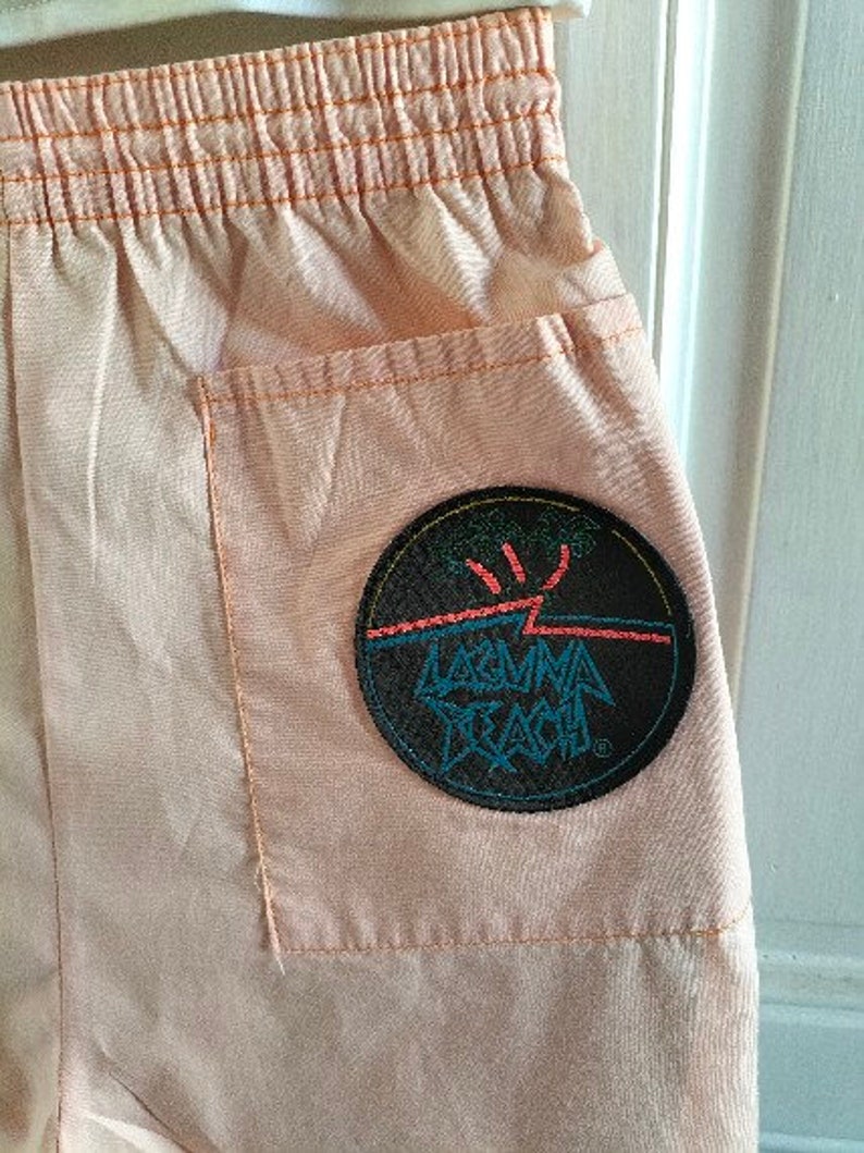 Puede incluir: Pantalones cortos de color melocot&oacute;n con un parche negro grande en el bolsillo trasero. El parche tiene un rayo blanco y el texto "Laguna Beach" en azul y rosa.