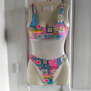 May include: A two-piece swimsuit with a patchwork design. The top is a scoop-neck style with wide straps. The bottoms are high-cut. The swimsuit features a mix of floral, gingham, and geometric patterns in pink, blue, yellow, and green.